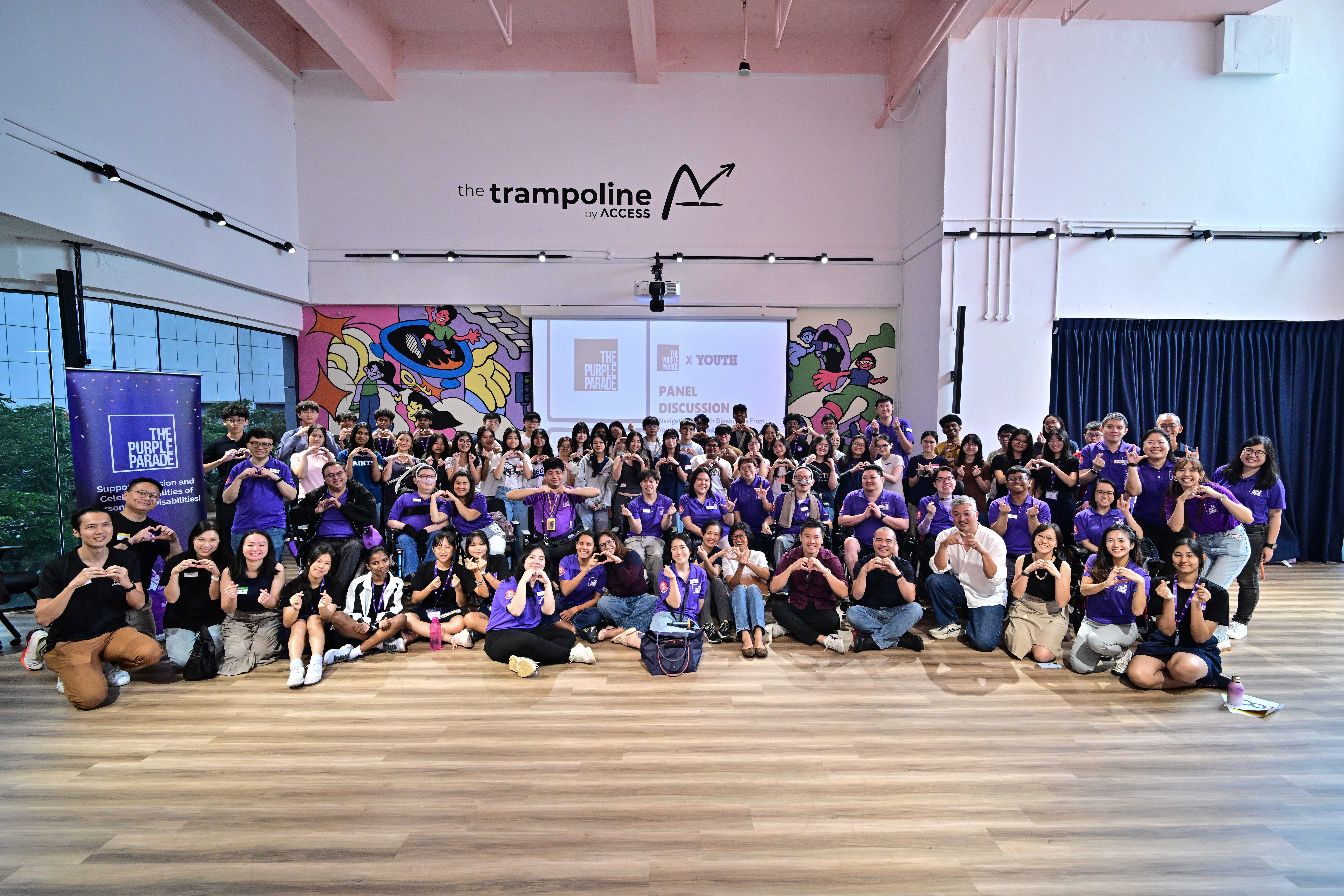 A large group of participants, many in purple T-shirts, make heart gestures at the Purple Parade Youth Conversations panel discussion held at The Trampoline by Access.